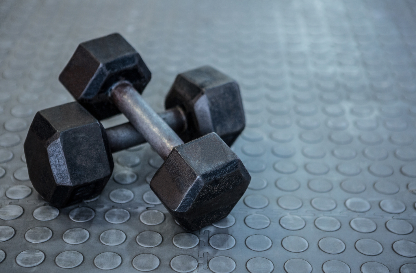 Two black metal dumbbells resting on a grey textured rubber gym floor with a pattern of small raised circles, showing the mat surface detail.