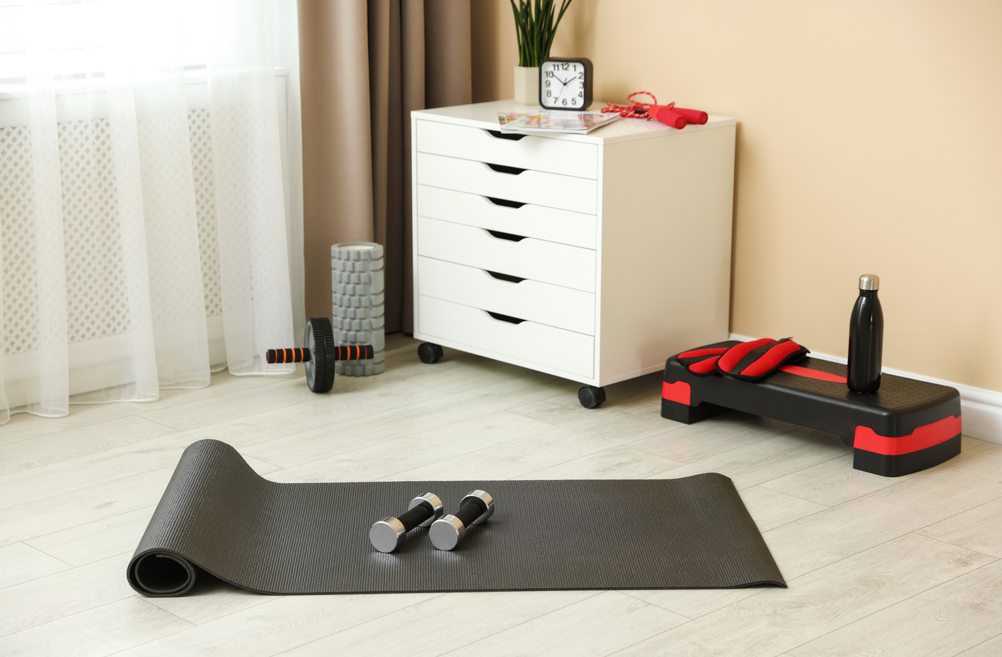 A white drawer cabinet on casters holds a jump rope and a clock, with a foam roller, an ab wheel and a step platform beside it and a mat with two dumbbells on the floor.