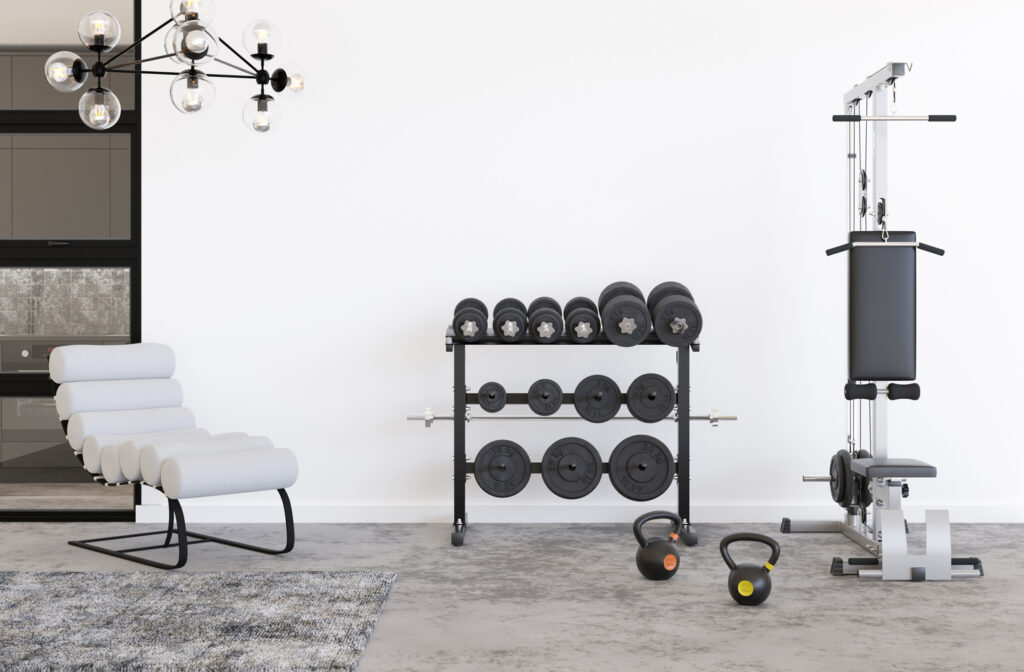 A bright home gym with a white chair, a multi-tiered rack of weights and dumbbells, and a weight machine standing on a gray floor, with two kettlebells in front of the rack.