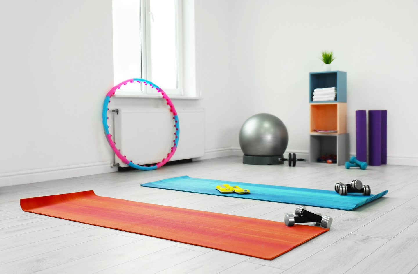 An orange yoga mat on a light floor in front of a blue mat holding exercise equipment, with a hula hoop, a gray exercise ball and a small colorful shelf of workout accessories in a room with a white wall and a window.